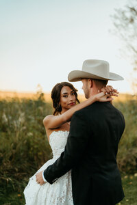 Bride in wedding dress holding groom wearing cowboy hat during outdoor country wedding portrait session.