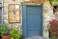 Framed painting hung on a stone wall beside a weathered blue door in a European village