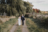 Couple de mariés marchant main dans la main dans un sentier champêtre au Vignoble de la Bauge, en pleine nature automnale, capturé par Jeff Gagnon photographe.