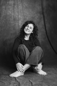 A black and white picture of a woman with dark curly hair sitting cross-legged on a dark gray backdrop. She wears jeans and a black long-sleeve shirt. She leans back with her hands on her feet as she smiles brightly, eyes closed, towards the camera.