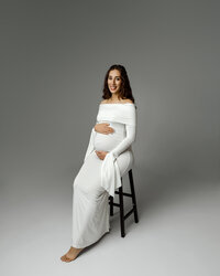 Pregnant woman smiling while sitting on a stool in a long white dress, gently holding her baby bump during a Yucaipa maternity photoshoot with Kaitlyn Dawn Photography.