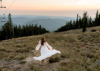 oregon city high school senior dancing in her white flowy dress in an open field at timberline lodge on mount hood with a view of the oregon cascade mountains behind her.