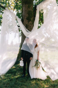 bride and groom standing in front of a tree draped in fabric at their Ranch Wedding