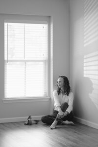 Person seated on the floor in the corner of an empty white room, sunlight falling through window blinds.