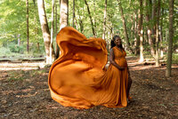 A expecting woman in a flowing orange gown poses in a wooded area, the fabric dramatically lifting in the air behind her.