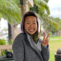 a young Asian woman wearing a hooded grey top is looking and smiling at the camera doing a peace sign with her hand