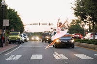 A ballerina in a pink tutu leaps through a crosswalk with car headlights alight behind her.