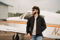 Senior portrait at a small hangar near Mobile International Airport in Mobile, Alabama — black
jacket and aviators in front of a small airplane, looking to the side.