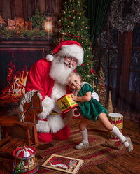 Young girl proudly shows Santa a favorite toy during a Santa's Magical Christmas photography session in Jacksonville, Florida, beside a glowing fireplace, vintage Christmas toys, and the cozy magic of the holiday season.