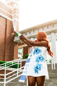 Senior popping champagne wearing white and blue flower dress as sunset on steps of TSU
