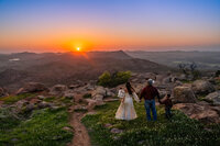 A family of four walks hand in hand toward a breathtaking mountain sunset, surrounded by rocky terrain and wildflowers atop the Wichita Mountains in Oklahoma. The mother wears a flowing dress and carries a baby, while the father and young son wear Western attire and cowboy hats.