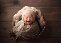 newborn baby girl wrapped in sage knit wearing a bear hat with bow in a basket. baby is smiling