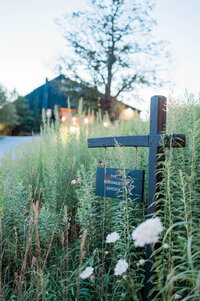 A wooden post holds a dark green sign reading “The Scribner’s Garden” amid tall wildflowers, with a warmly lit building in the distance.