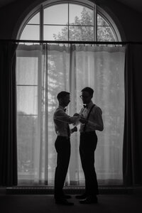 Moment intime et complice entre deux mariés LGBTQ lors des préparatifs, devant une grande fenêtre voilée dans les Laurentides — une scène élégante et authentique, capturée par un photographe de mariage à Montréal, célébrant la diversité, l’amour et la tendresse de leur connexion.