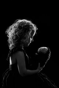 A black and white picture of a small blonde girl holding a stuffed otter. They are artfully lit against a black background.