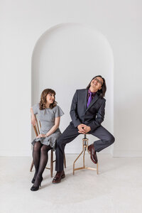 A woman in a gray dress smiles at a man in a dark suit and purple tie, playfully seated sideways on a stool—an editorial portrait photography Washington DC metro session captured by a white arched wall.
