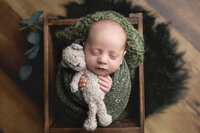 Newborn boy wrapped in a hand-knitted soft green wrap snuggling a bear during his newborn session at Jennifer Brandes Photography in South Central, Minnesota.