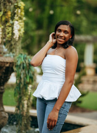 Senior portrait at Bella Sera Gardens in Loxley, Alabama — standing in the courtyard holding
her hair, wearing a white top and blue jeans.