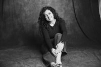 A black and white picture of a woman with dark curly hair sitting on a dark gray backdrop. She wears jeans a black long-sleeve shirt. One leg extends out in front of her as the other is bent upward with her arms softly wrapping it. She smiles brightly and tilts her head to one side.