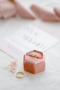 A pink ring box with a gold wedding band sits on white linen fabric in a close-up product photography shot.