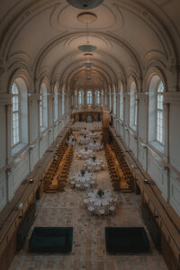 Vue en hauteur de la salle de réception voûtée à l'Abbaye d'Oka, tables rondes dressées en enfilade, arches et fenêtres cintrées, ambiance intime en fin de journée