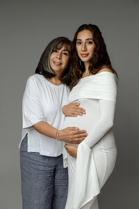 Pregnant woman in a white gown standing with her mother, both gently holding her belly during a maternity session by Kaitlyn Dawn Photography in Yucaipa.