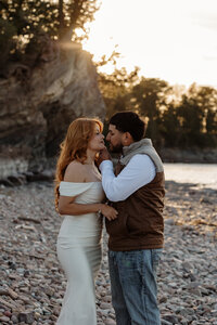 A couple stands by the water at sunset, leaning in for a kiss in New England. Fine art engagement photography by Damini Photography, capturing the golden light, romantic tension, and intimate connection of the moment.