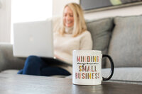 A close-up of a white mug that reads “Minding my own small business” with a blurred female entrepreneur working on her laptop in the background—capturing the essence of small business life.
