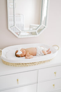 Newborn baby girl with white bow lays on changing table during in-home newborn session