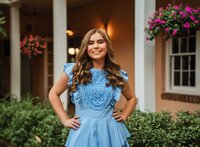 Senior portrait at the Malaga Inn in Mobile, Alabama— wearing a blue dress in the courtyard, hands on hips, with greenery and pink flowers in the background.