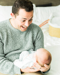 Father smiling and looking down at his newborn baby during a candid in-home lifestyle newborn photography session.