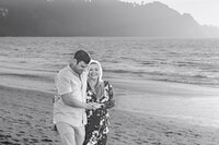 Black and white engagement photo of a couple walking toward the camera on the beach at sunset.