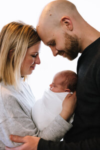 A backlit picture of two parents snuggling their newborn daughter. She is wrapped in white lying between them and the parents nuzzle their heads together.