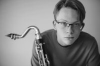 Black and white portrait of a young musician in glasses holding a saxophone against a plain backdrop.