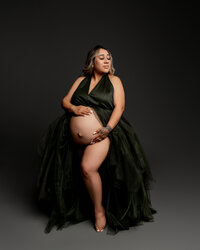 Artistic maternity photography portrait of pregnant woman seated wearing a dark green tulle maternity gown with dramatic lighting highlighting her baby bump by Yucaipa best maternity photographer Kaitlyn Dawn Photography.