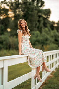 Senior portrait in downtown Magnolia Springs, Alabama — sitting on a white wooden fence with the sunset glowing behind her hair.