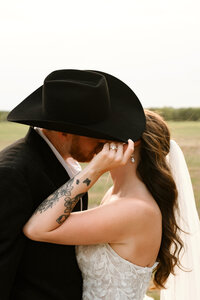 a bride and groom kiss behind a cowboy hat during their couples portraits at hidden jewel in blue ridge, texas.