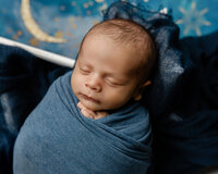 Sleeping newborn baby boy wrapped in a blue swaddle and posed against a dreamy moon and stars backdrop during a creative newborn photography session by Kaitlyn Dawn Photography.