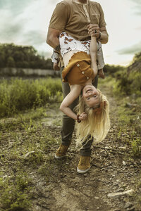 Girl with blonde hair wearing butterfly pants and tank top is held upside down by her dad while she smiles and flexes in outdoor family phtography