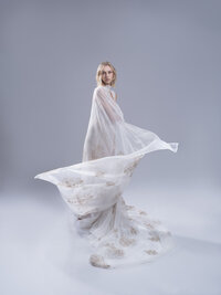 Model in a sheer white gown with the train fanned across the floor of a pale studio.