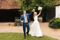 A newlywed couple joyfully walks outdoors on a lawn. The groom, in a blue suit, holds a drink in one hand and the bride’s hand with the other. The bride wears a white dress and holds a bouquet, both smiling and celebrating.