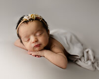 Peacefully sleeping newborn baby girl wearing a delicate floral headband and resting on a soft neutral backdrop during a studio newborn photography session by Kaitlyn Dawn Photography.