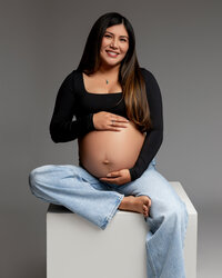 Pregnant woman sitting on a white studio cube wearing jeans and a black top during a maternity session by Kaitlyn Dawn Photography.