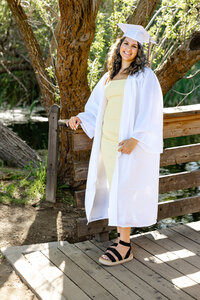 High school graduate wearing a white cap and gown standing on a wooden bridge during golden hour, graduation portrait by Kaitlyn Dawn Photography.