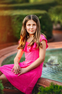 Young girl in pink dress sitting beside a stone fountain with warm golden light