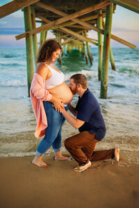 Expectant father kissing mother’s belly under the pier during an Atlantic Beach maternity session