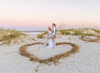 Professional photography by Nature's Child Photography: St. Augustine wedding capturing a playful beach moment as the bride holds her groom in a mock dip for a kiss, standing inside a heart formed in the sand beneath a pastel sky.