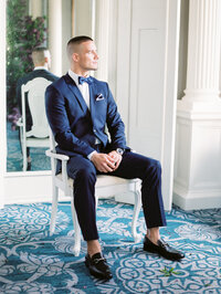 groom seated in a chair near a window wearing a navy suit in a refined editorial portrait