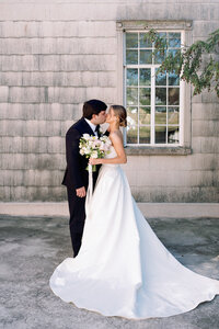 Bride and groom sharing a kiss while holding a bouquet in front of the stone wall at Burritt on the Mountain in Huntsville, Alabama, photographed by Perry Hancock.