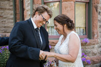 Two brides laughing and exchanging rings during their outdoor LGBTQ wedding ceremony at a historic stone venue in Colorado.